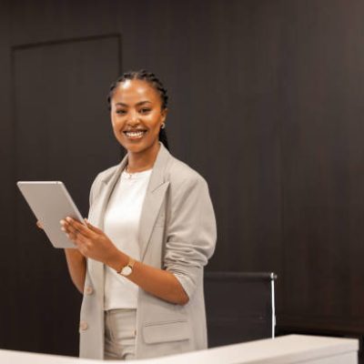 The young African-American female receptionist stands behind the reception desk with a digital tablet in her hands and smiles at the camera.