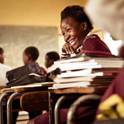 A Happy young South African girl (from the Xhosa tribe) works on her studies and jokes with her friends at at an old worn desk in a class room in the Transkei region of rural South Africa.
